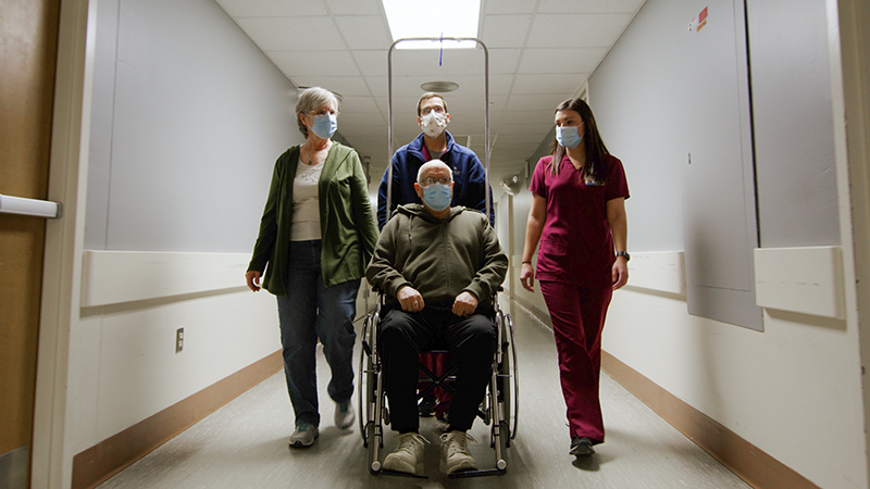 Three providers walking down hall, middle person pushing a patient in wheelchair