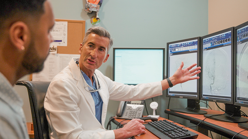 Male surgeon point to a bank of computer monitors, explaining something to a male patient