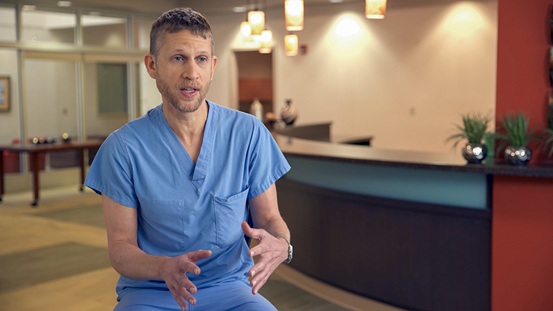 Provider in blue scrubs, seated in open area, talking and using his hands to explain something