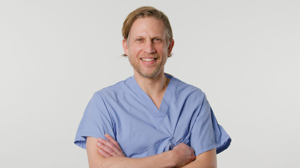 Male provider in blue scrubs, arms crossed, smiling at camera