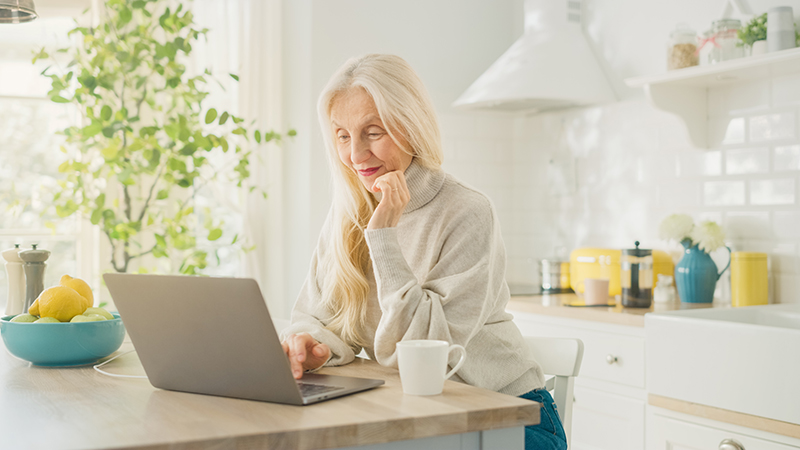 Older woman I her kitchen reading something on laptop computer